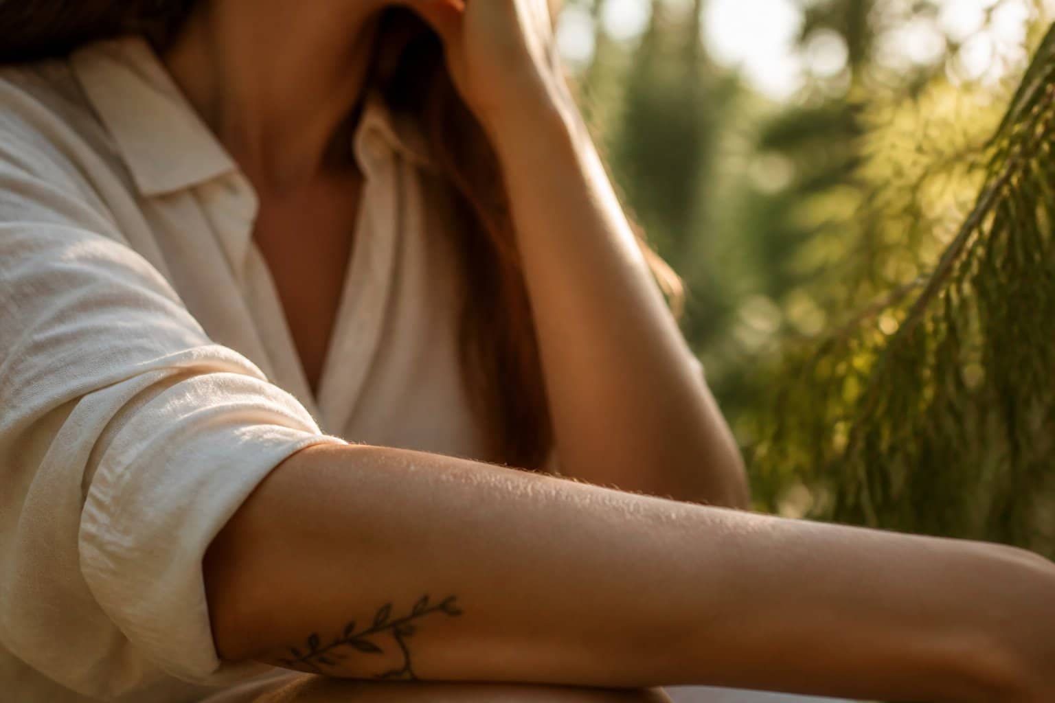 Braço de uma pessoa com uma tatuagem discreta, em um ambiente iluminado pela luz natural com folhagens ao fundo.