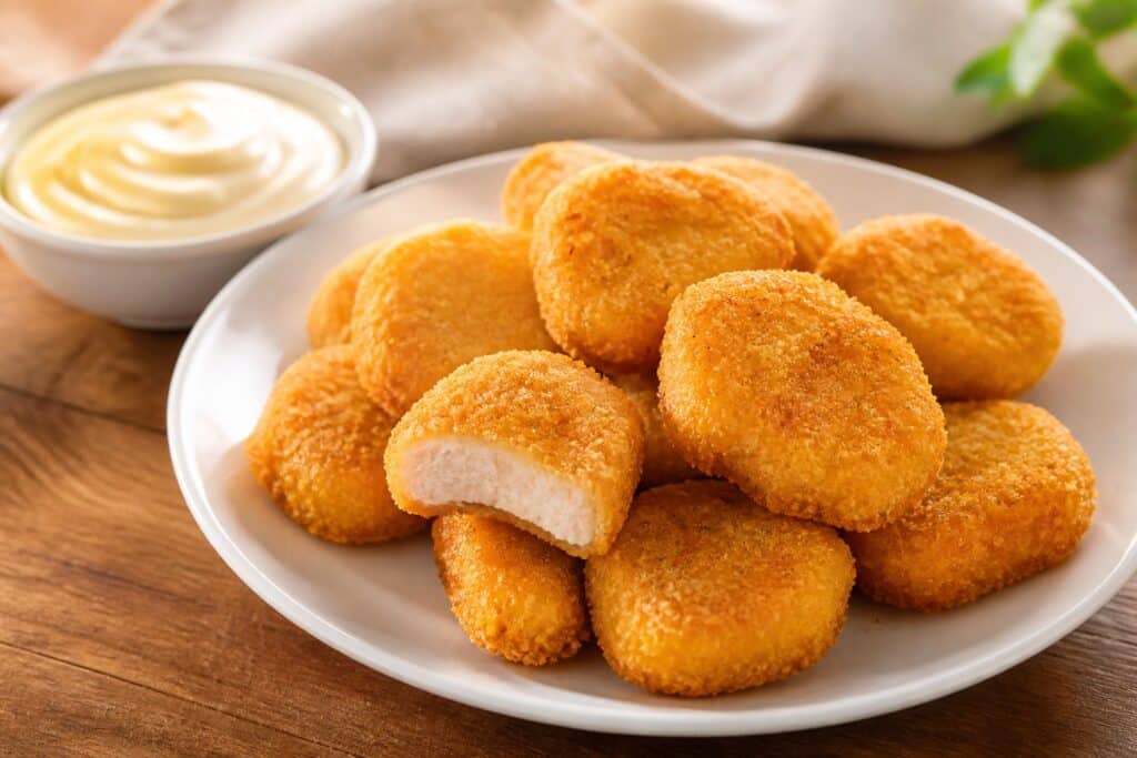 Prato com nuggets dourados e crocantes acompanhados de um pequeno recipiente com molho cremoso sobre uma mesa de madeira.