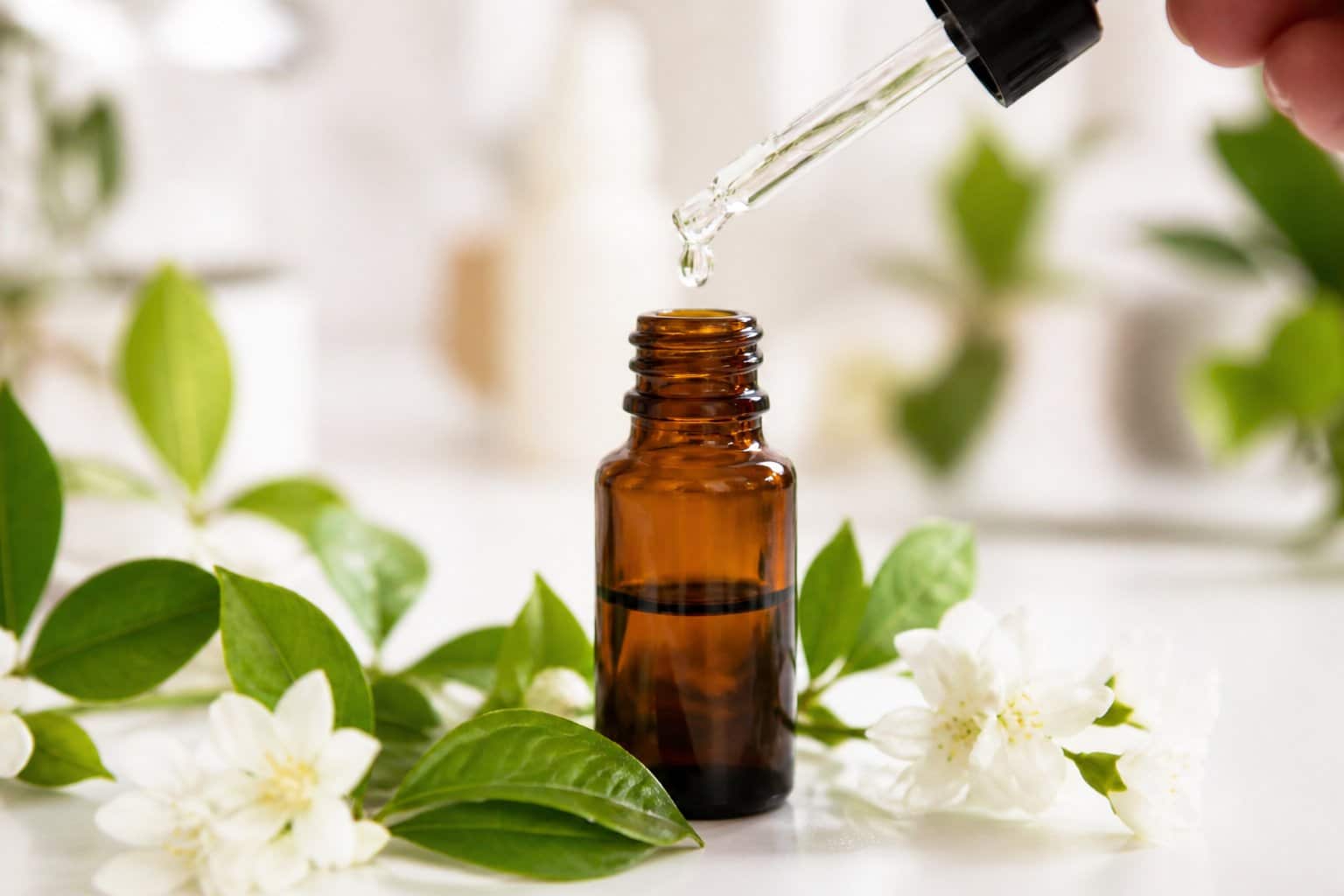 Frasco âmbar com conta-gotas pingando líquido transparente, cercado por folhas verdes e flores brancas sobre superfície branca.